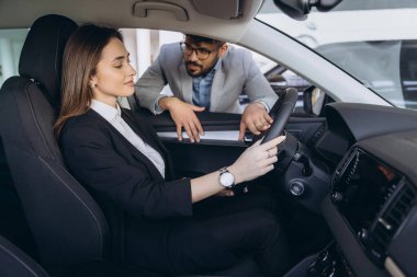 Salesperson explaining car functions to potential customer sitting at steering wheel in auto showroom