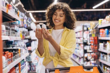 Young woman with curly hair comparing two deodorants while browsing the aisles of a supermarket, making a thoughtful shopping decision