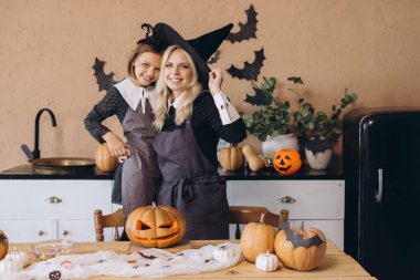 Mother wearing witch hat and daughter smiling and preparing Halloween pumpkins in kitchen with bats and candies