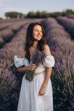 Smiling young woman in white dress holding a bouquet of freshly picked lavender in a beautiful lavender field at sunset