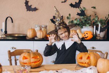 Girl wearing witch hat making a scary face with carved pumpkins in the foreground, celebrating Halloween