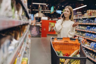 Markette alışveriş arabasıyla alışveriş yapan genç bir kadın telefonda konuşuyor.