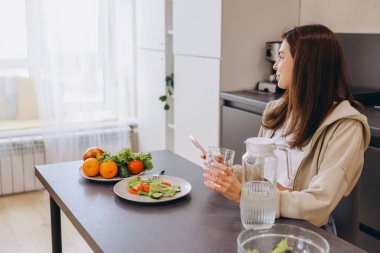 Young woman sitting at kitchen table, enjoying a healthy meal of salad and fresh fruit while browsing her smartphone