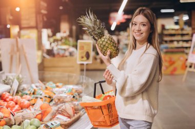Young woman joyfully holding a ripe pineapple while shopping for fresh groceries in a vibrant supermarket filled with healthy options