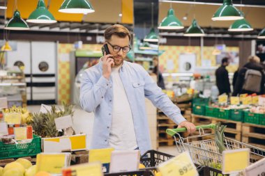 Customer using smartphone while pushing shopping cart in supermarket with fresh fruit and vegetables