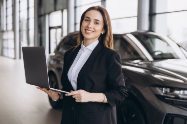 Saleswoman smiling and holding a laptop while posing in a car dealership with a new car in the background