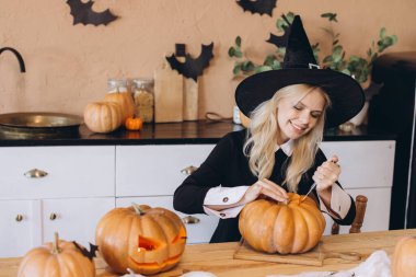 Smiling young woman wearing a witch hat, joyfully carving a pumpkin in the kitchen for a festive Halloween party celebration