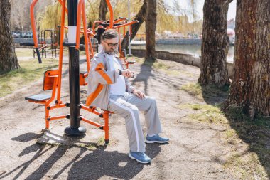 Bearded senior man checking his smartwatch while resting on outdoor gym equipment in a sunny park, enjoying a moment of relaxation