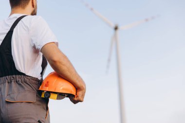 Technician holding an orange helmet, observing a wind turbine at a sustainable energy plant, contributing to renewable energy solutions