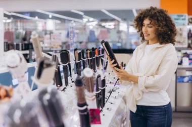 Young woman comparing hair straighteners, shopping for hair care appliances in a large electronics store