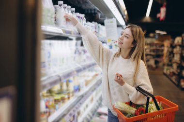 Genç bir kadın markette alışveriş yaparken süt şişesine uzanıyor, rafları taze yiyeceklerle dolduruyor.