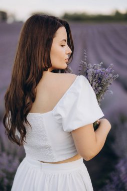 Woman dressed in a flowing white gown, holding a fragrant lavender bouquet while surrounded by vibrant purple blooms in a serene field
