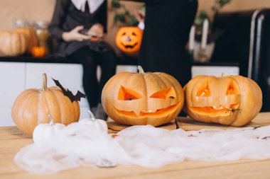 Three carved Halloween pumpkins on a table with a mother and daughter preparing for the holiday in background