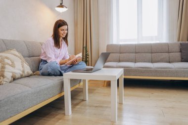 Young woman sitting on sofa working from home taking notes on notebook while watching her laptop placed on a small white table