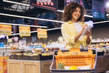 Smiling woman with curly hair checking her shopping list while pushing a shopping cart full of groceries in a supermarket