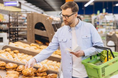 Man wearing disposable glove picking up pastry bun and holding grocery list while shopping for groceries in supermarket
