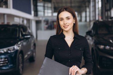 Saleswoman smiling and holding a clipboard while posing in a modern car dealership