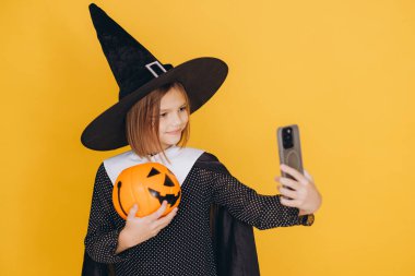 Smiling girl wearing witch costume holding carved pumpkin and taking selfie with her smartphone on yellow background