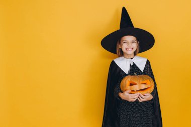 Little girl wearing Halloween witch costume holding carved pumpkin with spooky face smiling on yellow background