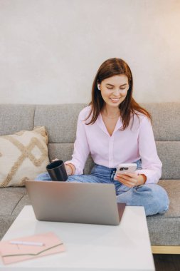 Smiling woman sitting on sofa with laptop and smartphone, enjoying a cup of coffee while working remotely