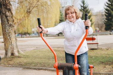 Elderly woman exercising on elliptical trainer in a park, promoting healthy lifestyle and active aging