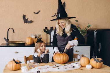 Mother carving a pumpkin while her daughter is looking at her, both wearing witch costumes, preparing for a Halloween party