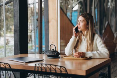 Young woman working remotely in a cozy cafe, sipping tea and chatting on her mobile phone while focused on her laptop