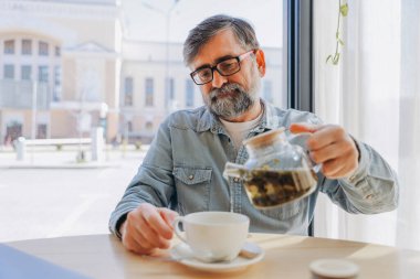Bearded senior man pouring herbal tea from a glass teapot into a white cup, enjoying a relaxing moment in a cafe