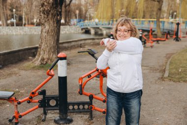 Happy senior woman stretching her arm at an outdoor gym in a park, enjoying a healthy and active lifestyle