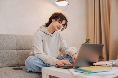Teen student girl sitting on the sofa is studying and using laptop for her homework, with books and a smartphone near her
