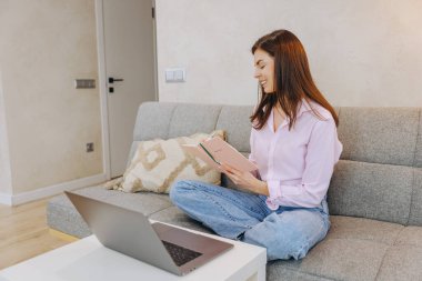 Smiling young woman sitting on couch writing notes in notebook, working remotely with laptop on coffee table in living room