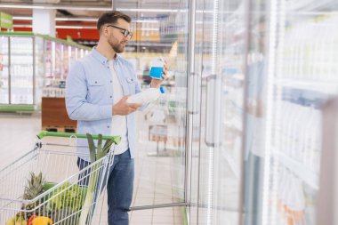 Man comparing two milk or yogurt bottles while shopping groceries in supermarket refrigerator aisle