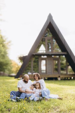 Happy family sitting together on lush grass in front of their charming a frame house, enjoying quality leisure time under the sun