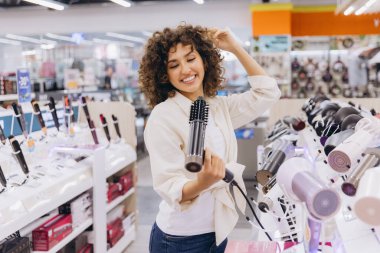 Smiling woman with curly hair testing a new hair dryer while styling her hair in a vibrant appliance store filled with modern products