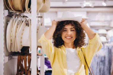 Smiling young woman with curly hair trying on a white hat in a clothing store, enjoying her shopping experience