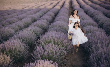 Mom and daughter wearing white dresses are walking through rows of lavender in bloom, enjoying a sunny summer day