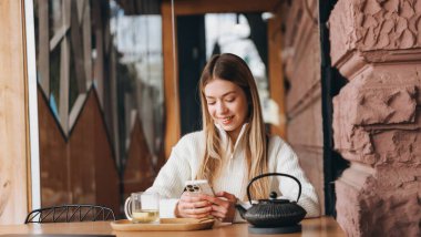 Smiling woman in a cozy cafe texting on her smartphone, with a cup of tea and teapot on the table, enjoying a relaxing moment