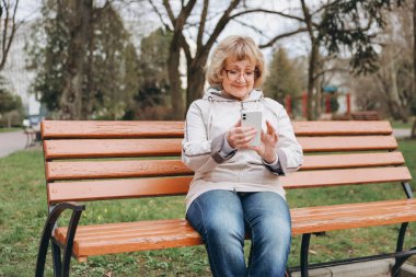 Elderly woman using smartphone while sitting on a bench in a park, enjoying mobile technology in nature
