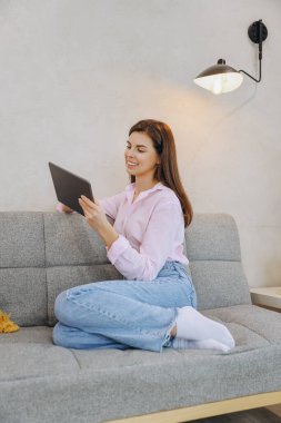 Young woman using a tablet and smiling while sitting comfortably on a sofa in her modern living room