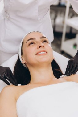 Professional beautician wearing black gloves applying cream on shoulder of smiling young woman lying on massage table in beauty salon