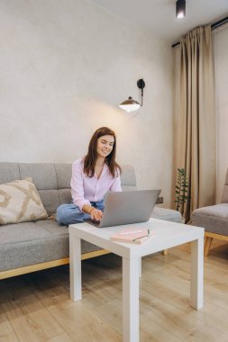 Smiling woman sitting comfortably on sofa using laptop with legs crossed working remotely from home