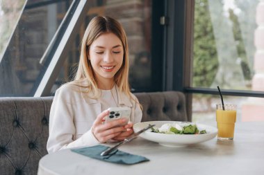 Businesswoman enjoying a nutritious meal while browsing her smartphone in a stylish restaurant, radiating happiness and relaxation