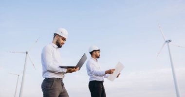 Two engineers are inspecting a wind turbine power plant, using a laptop and blueprints, with a clear blue sky in the background