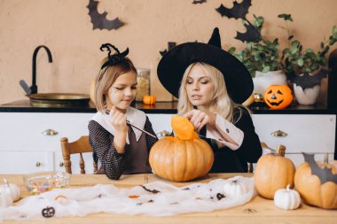 Mother wearing a witch hat and daughter joyfully carving a pumpkin together for a festive Halloween party at home