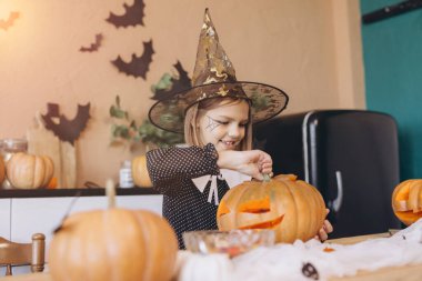 Smiling little girl in a witch hat joyfully carving a pumpkin, preparing for a festive Halloween party filled with fun and creativity