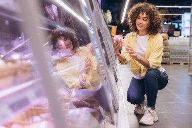 Smiling curly haired woman crouching and choosing frozen food from a refrigerated aisle in a brightly lit supermarket