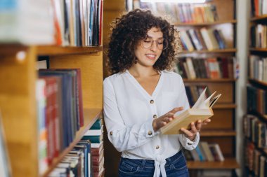 Curly haired student wearing glasses, standing next to a bookshelf, reading a book in the library, smiling