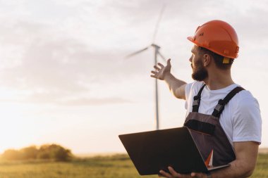 Engineer with a laptop explaining the operation of a wind turbine in a field during a beautiful sunset, highlighting renewable energy solutions