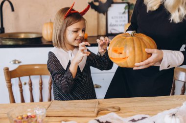Mother and daughter preparing a jack o' lantern from a pumpkin for Halloween, wearing costumes and having fun in their kitchen
