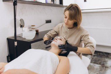 Beautician wearing black gloves applying a face mask to a client lying on a massage table in a beauty salon